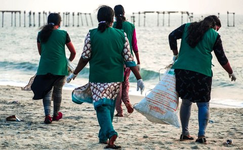 We see the backs of four women on a beach in green vests. Two of them are carrying sacks and all of them are wearing gloves. One woman is bending down to pick up a piece of plastic waste.