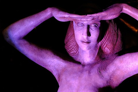 The photo shows the bust of a statue of a woman with her hands on her forehead looking out into the distance. It is lit in purple.