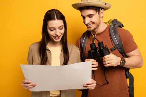 Auf dem Bild sind eine junge Frau und ein junger Mann zu sehen. Sie schauen beide auf eine Landkarte und lächeln. Er hält ein Fernglas.