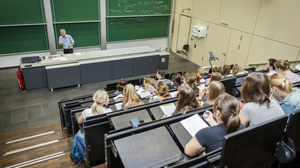 Studierende sitzen im Hörsaal bei einer Chemievorlesung. Professor steht an der Tafel. 