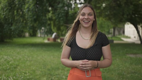 eine junge Frau steht lächelnd vor einem begrünten Hof mit Bäumen. Sie hat lange, offene blonde Haare, trägt eine orange Hose mit breitem Görtel, einem gepunkteten schwarzen Oberteil und einer goldenen Kette um den Hals. Ihre Hände sind verschränkt.