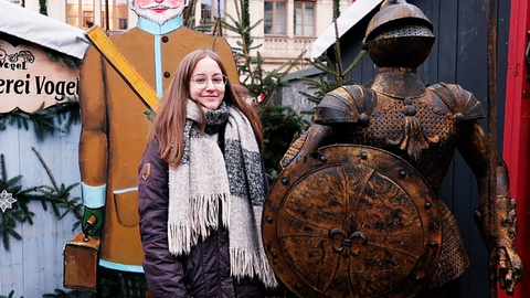 Foto: eine junge Frau mit langem, braunem Haar und Brille steht lächelnd zwischen einer Pappfigur und einer Ritterrüstung. Es ist weihnachtlich geschmückt.