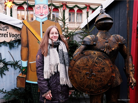 Foto: eine junge Frau mit langem, braunem Haar und Brille steht lächelnd zwischen einer Pappfigur und einer Ritterrüstung. Es ist weihnachtlich geschmückt.