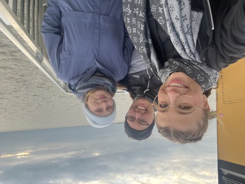 drei junge Frauen in warmer Kleidung stehen auf einem Boot und lächeln in die Kamera. Im Hintergrund ist das Meer zu sehen.