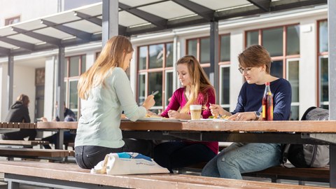 Drei junge Frauen sitzen beim Essen an einem Tisch im Außenbereich der Mensa.