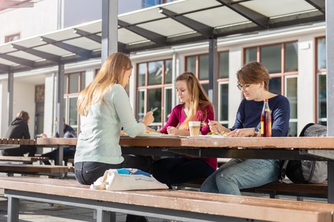 Drei junge Frauen sitzen beim Essen an einem Tisch im Außenbereich der Mensa.