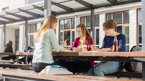 Drei junge Frauen sitzen beim Essen an einem Tisch im Außenbereich der Mensa.