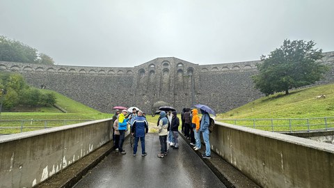 Foto von einer Gruppe Menschen mit Regenschirmen auf einer Straße, die direkt auf einen großen Staudamm im Hintergrund zulaufen. Links und rechts Wiese und Bäume.