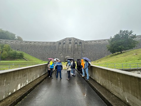 Foto von einer Gruppe Menschen mit Regenschirmen auf einer Straße, die direkt auf einen großen Staudamm im Hintergrund zulaufen. Links und rechts Wiese und Bäume.