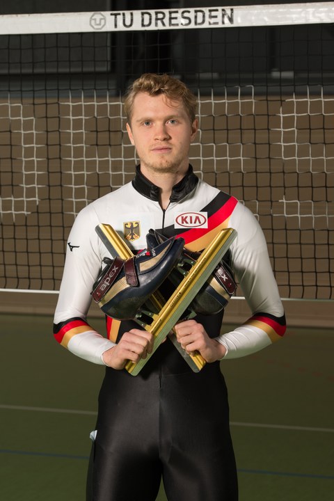 Foto mit Leon Kaufmann-Ludwig, Sportstipendiat 2019. Er steht im Deutschland-Sportdress mit vor der Brust gehaltenen Schlittschuhen in der Turnhalle vor dem Volleyball-Netz.