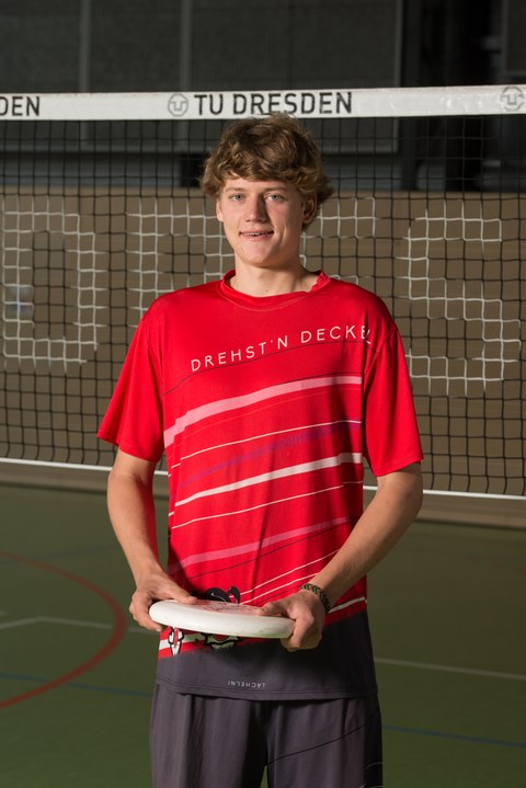 Foto mit Jakob Dieckmann, Sportstipendiat 2019. Er steht im Sportdress mit einem Frisbee in der Hand vor dem Volleyballnetz in der Turnhalle