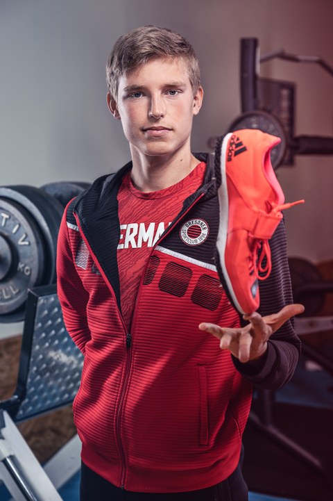 Porträt Jonas Wagner. Der Sportler in roter Trainingsjacke balanciert einen roten Sportschuh mit Spikes auf dem Zeigefinger. Er steht in einem Fitnessraum.