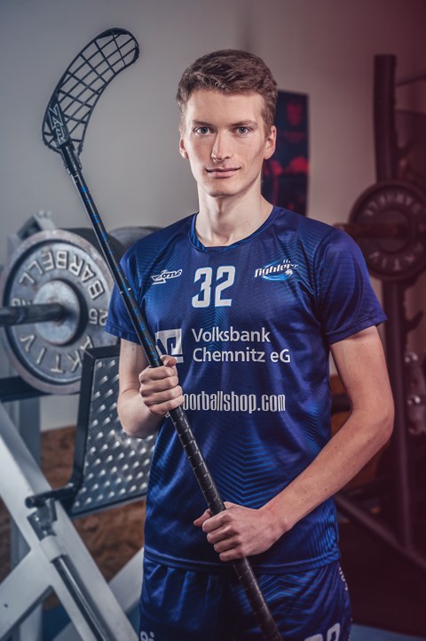 Porträt Gustav Peters. Der Sportler in blauem Wettkampf-Dress hat einen Hockeyschläger in der Hand und steht in einem Fitnessraum.