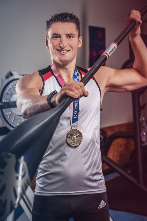 Porträt des Kanu-Sportlers Tom Liebscher. Er hat ein Paddel in der Hand, ist im weißen Sportdress, um den Hals hat er eine Olympia-Goldmedaille mit dem Schriftzug Tokyo 2020 auf blau, weißem Band hängen. Im Hintergrund ist ein Fitness-Raum zu erkennen.