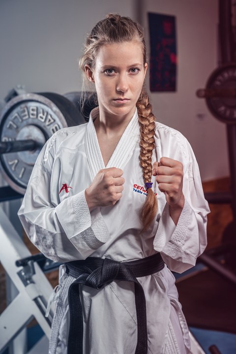 Porträt von Selina Stamer. Sie trägt eine weiße Karate-Uniform mit schwarzem Gürtel, hat die Hände in Brusthöhe zu Fäusten geballt und schaut herausfordernd in die Kamera. Ihre Haare hängen in einem eng geflochtenem Zopf über die linke Schulter.