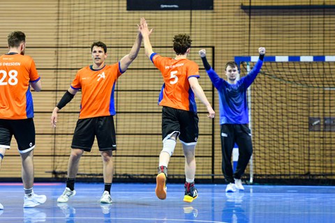 Wir sehen 4 Männer in sportlichen Outfits vor einem Handballtor in einer Halle, die beiden in der Mitte klatschen sich ab. Der Mann im Tor hebt seine Arme und ballt die Fäuste.