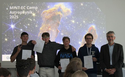 Vier Jugendliche und Prof. kobel stehen nebeneinander vor einem Hintergrund der "die Säulen der Schöpfung" zeigt mit der Aufschrift MINT-EC CAmp Astrophysik 2025". Die Jugendlichen halten Teilnahmeurkunden.