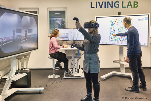 Das Foto zeigt eine blonde junge Frau die eine VR-Brille trägt und sich etwas anschaut. Im Hintergrund ist ein Mann der auf einem Bildschirm tippt und eine Frau in einem rosa Pullover die am Computer sitzt.