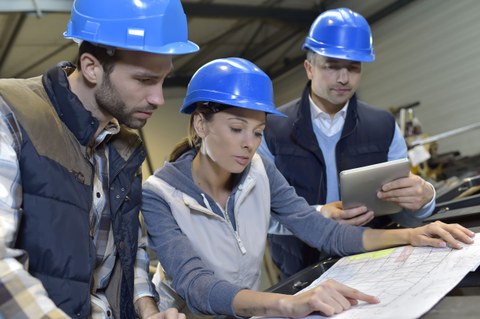Das Foto zeigt drei Personen mit blauen Helmen, die gemeinsam auf einen Bauplan schauen.