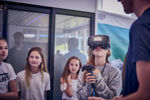 Das Foto zeigt junge Schülerinnen. Eine von ihnen hat eine VR-Brille auf und navigiert sich durch einen virtuellen Raum.