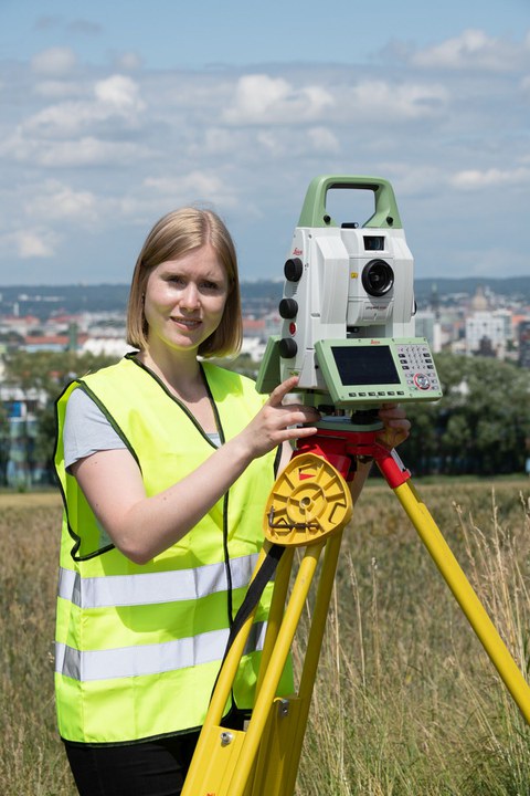 Das Foto zeigt eine junge blonde Frau die auf einer Wiese hinter einem Vermessungsgerät steht. Im Hintergrund ist die Stadt Dresden zu sehen.