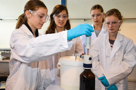 Das Foto zeigt vier junge Schülerinnen in Laborkitteln mit Schutzbrille, die in neugierig auf eine Apparatur schauen, die in einem Eimer etwas misst.