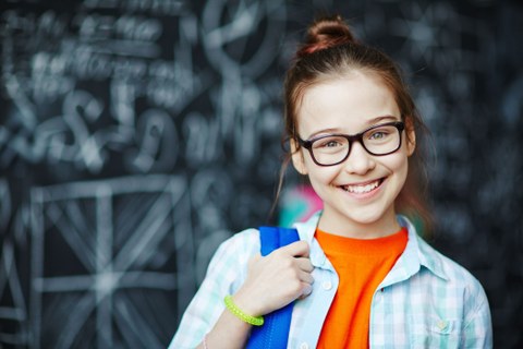 Das Foto zeigt ein junges Mädchen in orangem T-Shirt und Brille mit Rucksack über der Schulter, das in die Kamera lacht.