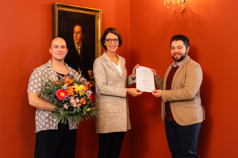 Hr. Saysaa steht neben Fr. Prof. Böhm, beide halten die Urkunde für den DAAD Preis. Hr. Schurig steht neben Frau Prof. Böhm und hält den Blumenstrauß.