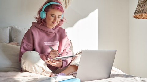 Ein Mädchen, das vor ihrem Laptop auf dem Bett sitzt und sich Notizen auf ihrem Tablet macht.