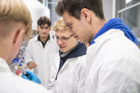 Vier junge Männer schauen auf ein kleines Reagenzglas. Sie tragen Laborkittel.