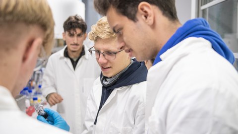 Vier junge Männer schauen auf ein kleines Reagenzglas. Sie tragen Laborkittel.
