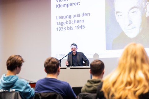 Im Vordergrund sind junge Menschen von hinten zu sehen. Vor Ihnen sitzt auf einer Bühne ein Mann am Mikrofon und spricht. Hinter ihm ist eine Folie mit Portrait Victor Klemperers und dem Titel der Tagebücher angeworfen.
