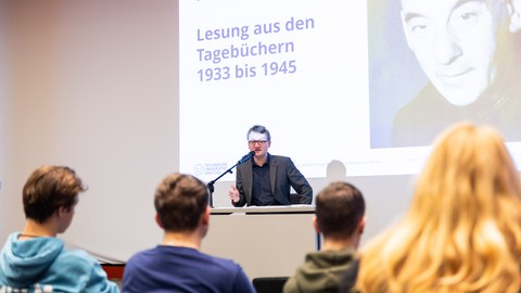 Im Vordergrund sind junge Menschen von hinten zu sehen. Vor Ihnen sitzt auf einer Bühne ein Mann am Mikrofon und spricht. Hinter ihm ist eine Folie mit Portrait Victor Klemperers und dem Titel der Tagebücher angeworfen.