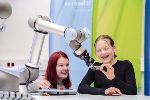 Ein Mädchen gibt einem Roboterarm einen Bauklotz. Daneben sitztz ein anderes Mädchen. Beide lachen.
