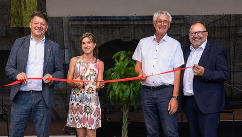 Vier Menschen halten ein rotes Band als Symbol der Eröffnung des Regionalbüros. An zweiter Stelle von links steht eine Frau, die mit einer Schere andeutet, das Band zu schneiden. Die anderen drei Menschen sind Männer. Alle sind festlich gekleidet.