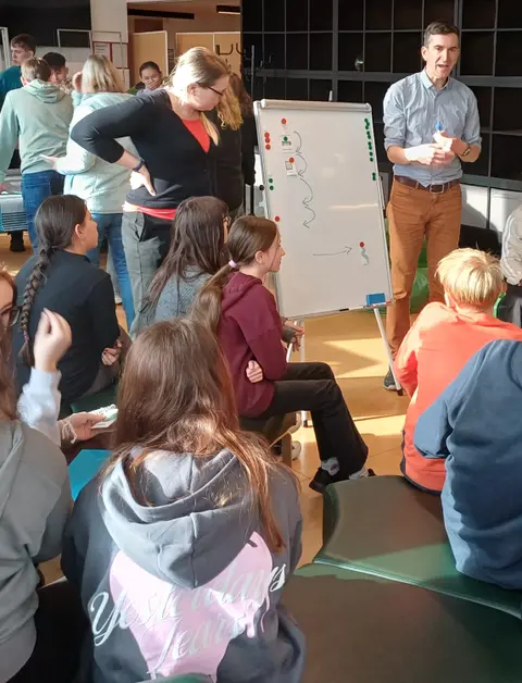 Jugendliche sitzen auf Sofas und hören einem Mann und einer Frau zu, die etwas auf ein Whiteboard zeichnen.