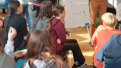 Jugendliche sitzen auf Sofas und hören einem Mann und einer Frau zu, die etwas auf ein Whiteboard zeichnen.