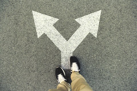 Auf dem Foto sieht man einen Doppelpfeil auf einer asphaltierten Straße. Die Pfeilspitzen zeigen nach rechts und links. Auf dem Pfeil steht eine Person, deren Beine und Füße zu sehen sind.