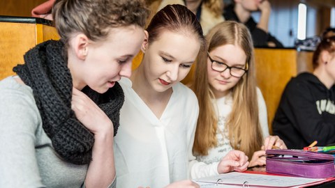 Photo of three female students sitting in a lecture hall and looking at written sheets in a red folder.