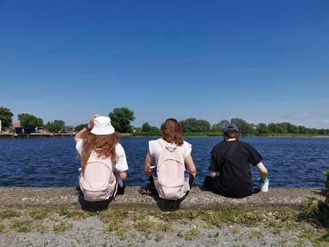 Drei Jugendliche sind von hinten zu sehen, die auf einer Mauer an einem See sitzen. Die obere Bildhälfte ist der blaue Himmel.