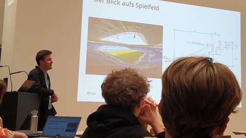 Zwei Schüler sitzen im Vordergrund, vor einer Präsentation über ein Stadiondach