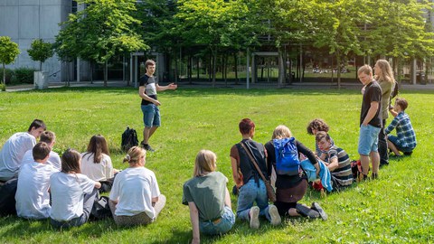 Foto. Eine Gruppe von Personen sitzt auf einer Wiese. Im Hintergrund ist ein Gebäude zu sehen, vor dem Bäume stehen. 
