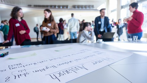 Menschen unterhalten sich in Kleingruppen, im Vordergrund ist ein Flipchartpapier mit der Aufschrift "interner Austausch" zu sehen