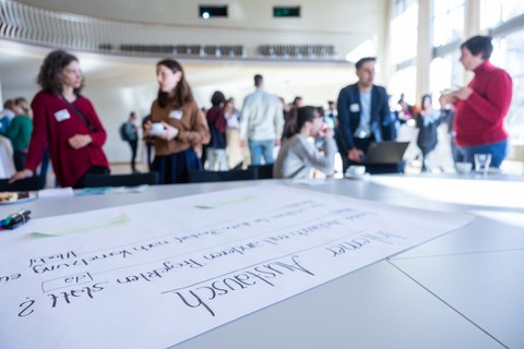 Menschen unterhalten sich in Kleingruppen, im Vordergrund ist ein Flipchartpapier mit der Aufschrift "interner Austausch" zu sehen