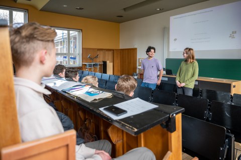 Zwei Schüler:innen stehen in Hörsaal und berichten sitzenden Schüler:innen.