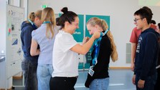 Eine Frau hängt einer Jugendlichen ein Band mit einem kleinen Schild um. Im Hintergrund stehen weitere Jugendliche.
