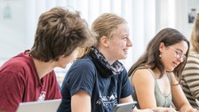 Jugendliche sitzen an einem Tisch in einer Reihe und sehen gut gelaunt aus.