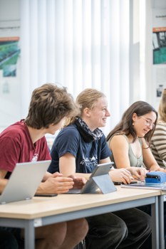 Jugendliche sitzen an einem Tisch in einer Reihe und sehen gut gelaunt aus.