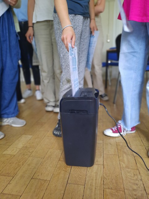 Ein Blatt wird in einem Datenschredder eingeführt. Im Hintergrund sind die Beine von vielen Personen zu sehen.