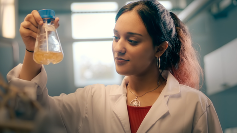 Eine junge Frau im Laborkittel betrachtet einen Glasbehälter mit einer Flüssigkeit.